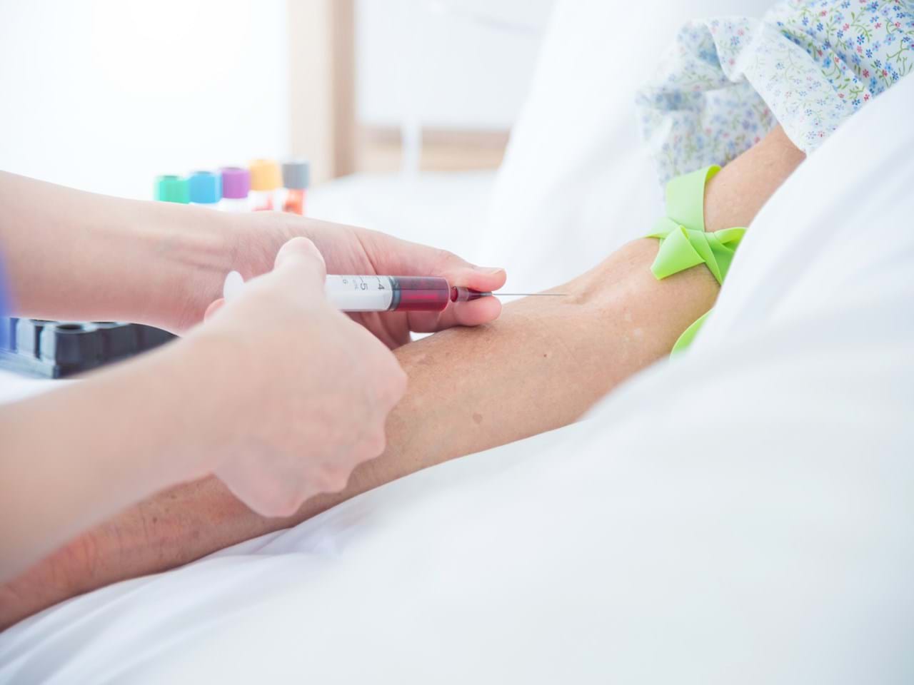 Eldre pasient får tatt blodprøve i armen av helsepersonell med grønt stasebånd.