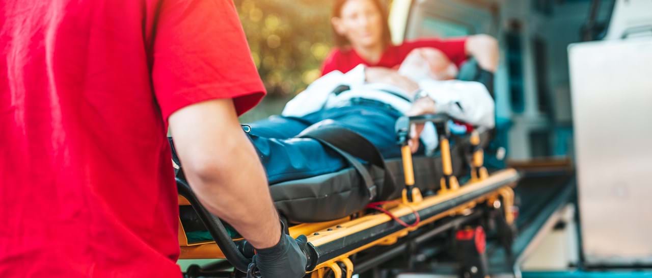 To ambulansearbeidere i røde uniformer laster en pasient på båre inn i en ambulanse. Pasienten er tildekket med et teppe og ser ut til å være bevisstløs.